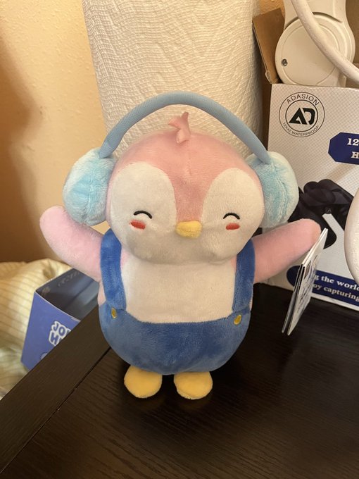 A small plush toy of a pink and white bird-like character wearing blue overalls and large blue headphones, sitting on a dark surface with packaging and what appears to be headphones visible in the background.