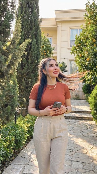 A young woman with long brown hair wearing a rust-colored top and beige pants stands outdoors near a beige house with cypress trees and citrus trees.