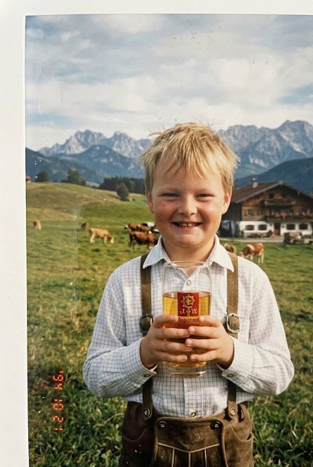A young blonde boy smiles while holding a glass mug of beer, dressed in traditional Bavarian lederhosen with suspenders, set against an alpine backdrop of cows and mountains—a playful self-portrait humorously suggesting the youthful "operator" behind the account. The date stamp "14 09 2004" is visible in the corner.