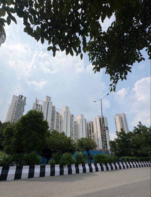 A photograph of modern high-rise residential apartment buildings viewed through tree foliage, with a street and black-and-white striped barrier in the foreground under a partly cloudy blue sky.