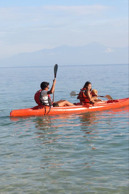 A man and a woman wearing life vests are kayaking together in an orange tandem kayak on calm water, with mountains visible in the hazy distance.