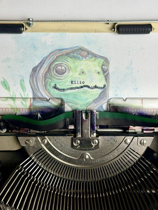 A typewriter with a paper featuring a hand-drawn, green frog-like character with a hood and a wide mouth full of teeth, labeled 'Kilio'.