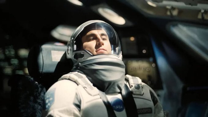 The image features a man wearing a spacesuit, possibly an astronaut, with his head resting against the seat of a vehicle or spaceship. There are two other people visible in the background, but they do not appear to be involved in the main subject's activity. The caption "Lock in" is written at the bottom right corner of the image.