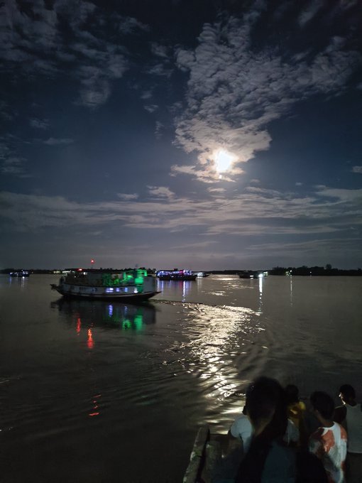 A nighttime riverside scene with a boat illuminated by green lights reflecting on the water under a moonlit cloudy sky, with people watching from the shore.
