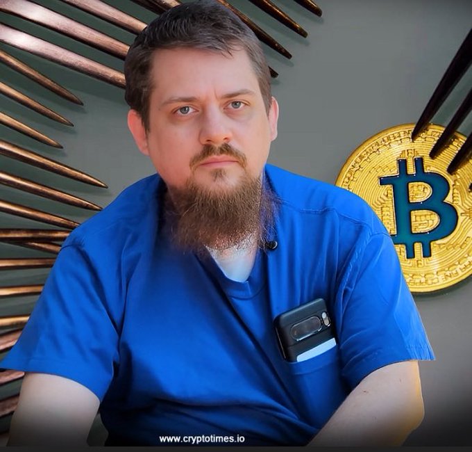 A bearded man in a blue scrub top sits in front of a decorative wall featuring a large Bitcoin symbol, with a 'www.cryptotimes.io' watermark at the bottom.