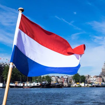 A Dutch flag waves prominently in the foreground against a bright blue sky, with Amsterdam's waterfront and historic buildings visible across the water. The image visually reinforces the post's news about the Netherlands repatriating 86 tonnes of gold to London, symbolizing the country's response to shifting global alliances and financial security concerns.