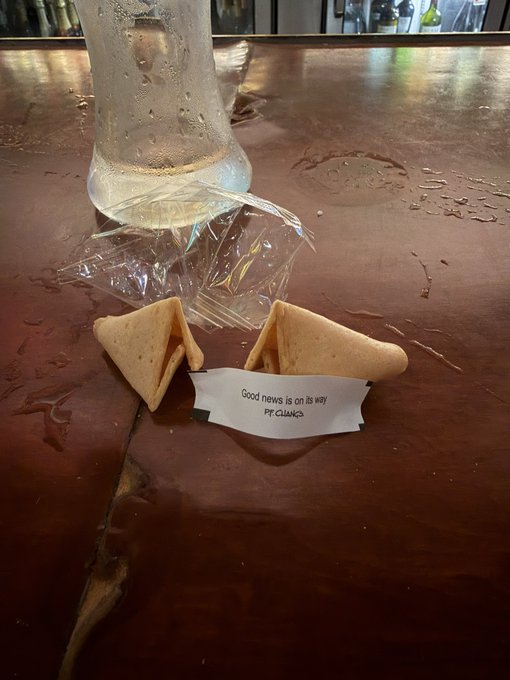 A photo of two fortune cookies on a bar counter with a slip reading 'Good news is on its way - PT CUANCA,' next to an inverted cup.