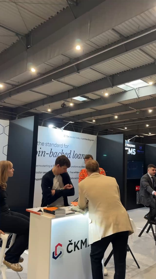 The image shows a group of people gathered around an exchange booth at a trade show, possibly attending or visiting a company exhibition. There are several individuals standing near the booth as well as some sitting on chairs nearby. The scene appears to be set up for an event with booths and displays showcasing different products and services.
