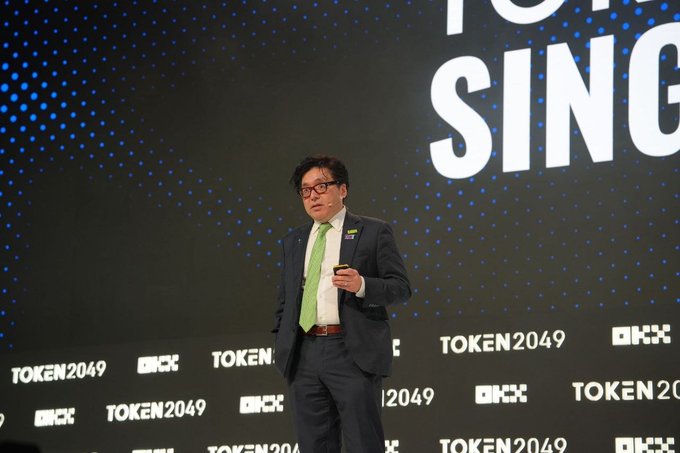 A man in a dark suit with a green tie speaks on stage at the TOKEN2049 conference, holding a device with the event's branding displayed on a large screen behind him.