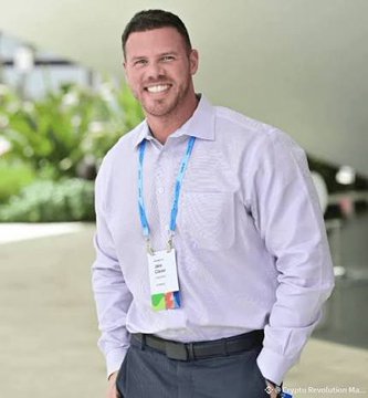 The image shows a smiling man in a light-colored button-up shirt wearing a lanyard with a badge, standing in what appears to be a conference or event setting. The post references Jake Claver (@beyond_broke) discussing patience with XRP holdings, but the photo does not depict Jake Claver—it appears to be a stock or unrelated image of a different attendee at a professional event.
