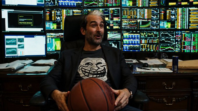 A man wearing a Trollface meme t-shirt sits holding a basketball in front of a wall of trading monitors displaying various stock charts.
