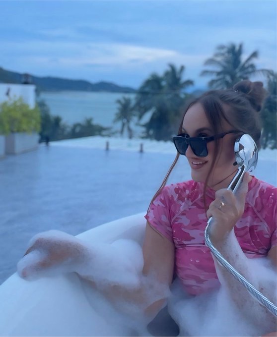 A woman in a pink camouflage shirt and sunglasses holds a showerhead while sitting in a bubble bath overlooking the ocean.