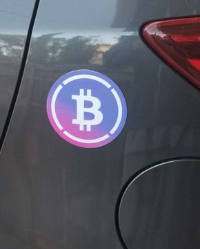 A circular purple and pink gradient sticker featuring the Bitcoin symbol is affixed to the rear of a gray car near its taillight.