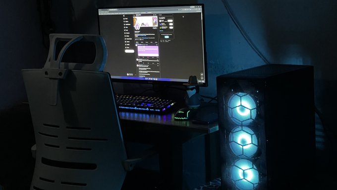 A dimly lit gaming setup features a curved monitor displaying a chat or stream interface, a mechanical keyboard with RGB backlighting, and a PC case with three glowing cyan fans.
