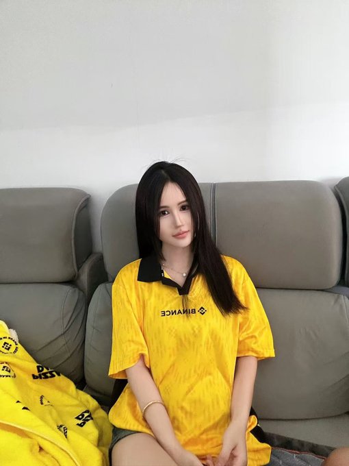 A young woman with long dark hair wearing a yellow Binance jersey sits on a gray couch.