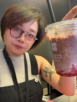 The image features a woman with short hair holding a drink, which appears to be a chocolate milkshake or smoothie from a shop called GM Morning Bloody drinks. She has a tattoo of a bird on her arm. The background shows a dining table where she is sitting and posing for the photo.