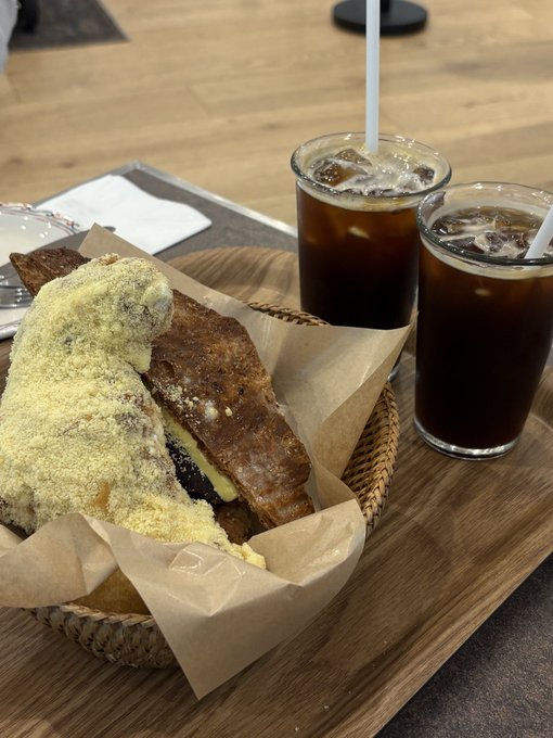 A woven basket holds a croissant and chocolate-stuffed pastry dusted with powdered sugar, accompanied by two iced coffees served with straws on a wooden tray. The casual café setting provides a relatable, everyday contrast to the high-stakes world of crypto investing mentioned in the post.