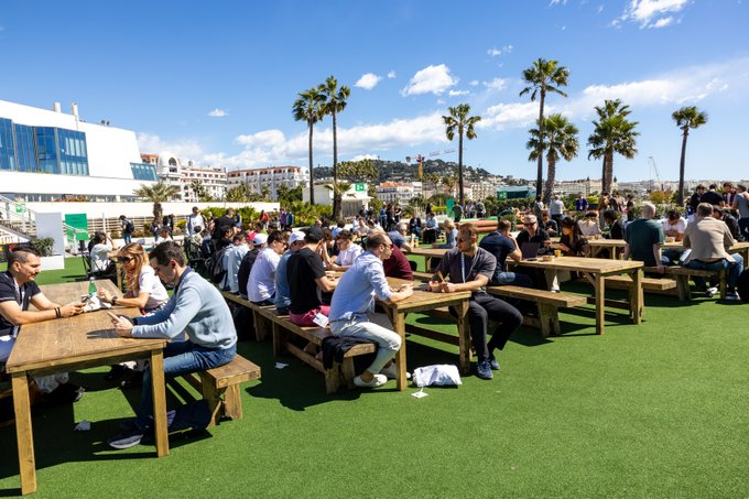 A sunny outdoor scene showing many people sitting at long wooden picnic tables on artificial grass, with palm trees and a coastal city view in the background.