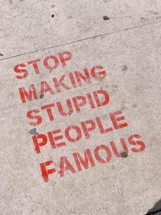 Red stenciled text on a concrete sidewalk reads 'STOP MAKING STUPID PEOPLE FAMOUS'.