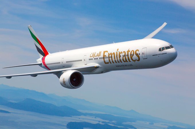 An Emirates Boeing 777-300ER is captured mid-flight against a backdrop of blue skies and distant mountains, with the airline's logo and Arabic script prominently displayed on its fuselage. The image visually represents the carrier now allowing UAE customers to book flights using Cryptocurrency.com Pay, tying the iconic airline to its new digital payment option.