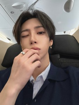 A selfie of a young man with dark hair styled down, wearing a dark jacket over a striped shirt, with his hand near his chin on what appears to be an airplane.