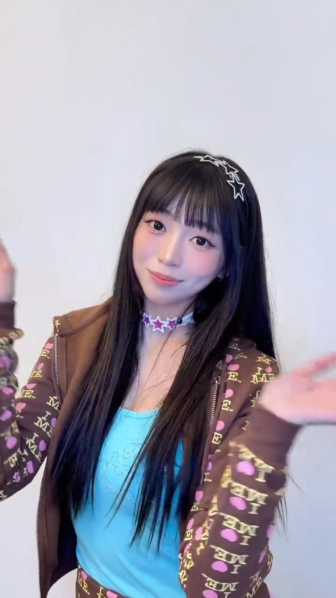 A young woman with long dark hair and bangs poses against a plain white wall. She wears a star-shaped hair clip, a matching star choker, a light blue tank top, and an open brown patterned zip-up hoodie, smiling slightly with one hand raised.
