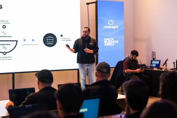 A man presents at the "Hack Buenos Aires" hackathon, with the "midnight" branding visible on a screen behind him, as developers work on laptops in the foreground. The scene captures the collaborative atmosphere of the event highlighted in the post, showcasing builders creating their first Midnight applications.