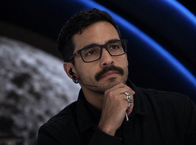 A man with glasses, a mustache, and a microphone earpiece rests his chin on his hand while wearing a black shirt and multiple rings. The image does not appear to relate to the post's instructions about acquiring Quotron tokens, claiming stock rewards, or buying wSPYx.