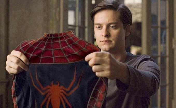 A scene from a Spider-Man film showing Tobey Maguire's character holding up his red and blue Spider-Man suit, revealing the iconic spider logo on the front.