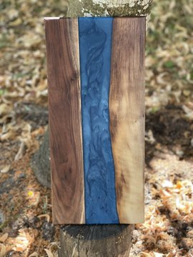 A handcrafted wooden vase or vessel featuring a striking blue resin river running through natural wood, displayed outdoors on a tree stump.