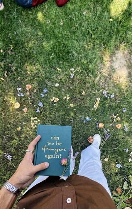 A hand holds a teal book titled "can we be strangers again?" amid scattered wildflowers on a sunny lawn, with white sneakers visible below. The romantic, contemplative scene pairs with the upbeat "gm web3 / happy monday frens" message, offering a reflective Monday mood among the crypto crowd.