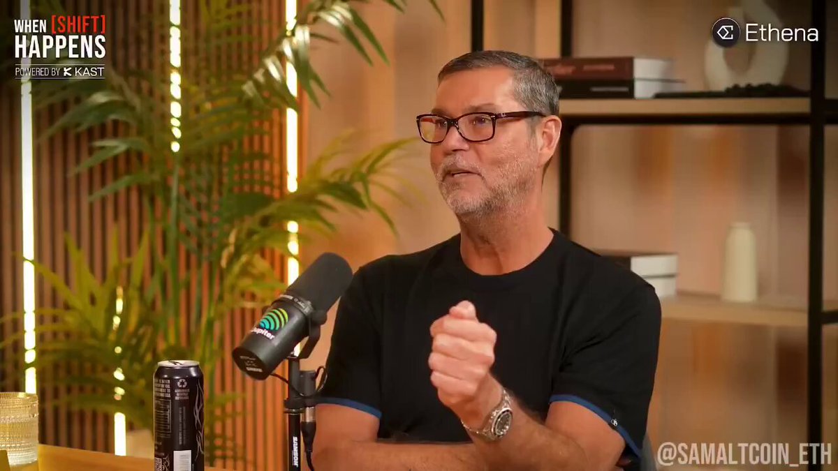 The image features a man wearing glasses, sitting at a desk with a microphone in front of him. He appears to be speaking into the microphone during an interview or discussion. The background includes a bookshelf filled with various items such as books and vases. There is also a potted plant near the bookshelf, adding some greenery to the scene.