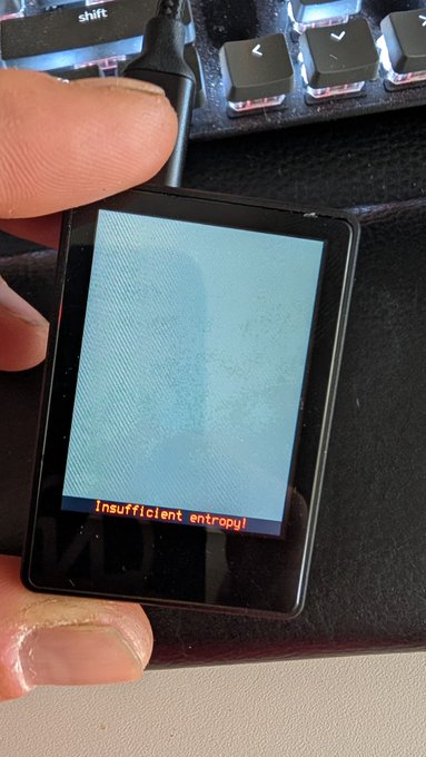 A hand holds a small device screen displaying the warning message "Insufficient entropy!" in red text on a light blue background, shown in front of a computer keyboard. This image illustrates the Krux wallet's entropy-checking feature, which alerts users when their seed generation lacks sufficient randomness for secure Bitcoin signing.