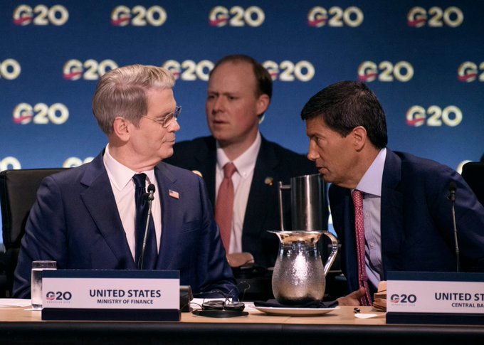 Two officials in suits converse at a G20 summit table with 'UNITED STATES MINISTRY OF FINANCE' and 'UNITED STATES CENTRAL BANK' nameplates visible against a backdrop covered in G20 logos.