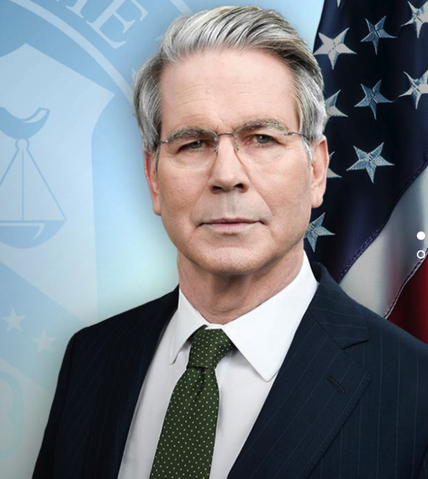 An official portrait of a gray-haired man in a dark pinstripe suit and green dotted tie, standing in front of an American flag and a Department of Justice seal.