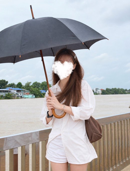 A woman in a white outfit holds a large umbrella along a riverside railing, with a cloudy gray sky overhead suggesting an approaching storm. The image visually reinforces the caption's stormy weather theme in Thailand, while the umbrella adds a playful, flirtatious metaphor for "sheltering" someone's heart.
