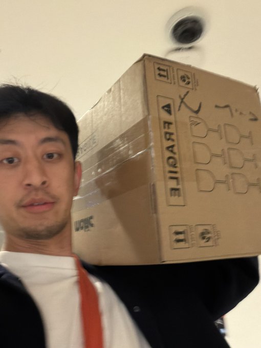 A person carries a large cardboard box marked “FRAGILE” and “UP,” posing indoors. The package is associated with @maticrobots, suggesting it contains one of their products.