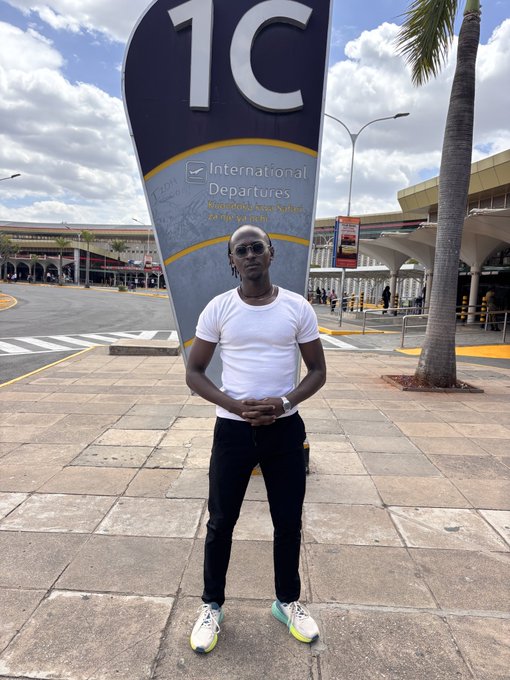 A tall man in a white t-shirt, black pants, and sunglasses stands in front of an 'International Departures' sign at an airport.