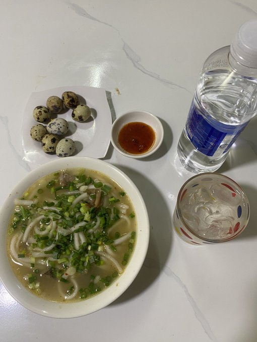 A meal on a white table featuring a bowl of noodle soup topped with scallions, a plate of quail eggs, a small dish of dipping sauce, a glass of ice water, and a bottle of water.