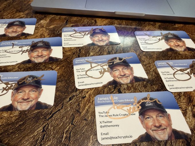 Business cards for "James Rule, Blockchain and Crypto Educator" are spread across a wooden surface beneath a laptop, featuring his social media handles (@allthemoney), YouTube channel "The James Rule Crypto Show," and email (james@teachcrypto.io), relating to the post by suggesting the bonus prize for winners includes personalized promotional materials like these custom cards.