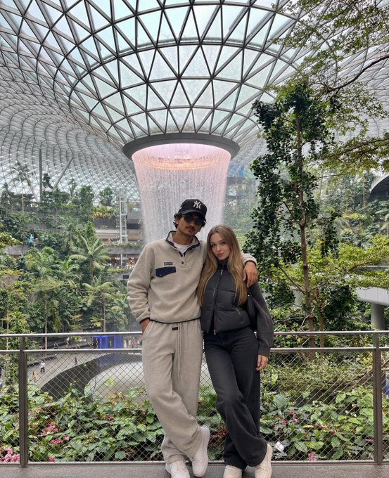A couple poses in front of the iconic Rain Vortex waterfall at Jewel Changi Airport, Singapore, wearing casual streetwear with one sporting a NY Yankees cap. The scenic indoor setting with lush greenery and the stunning glass-domed architecture serves as a perfect backdrop for attendees of the Token2049 crypto conference happening in the city.