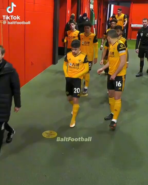 The image shows a group of people walking down an aisle, likely in a sports stadium or arena setting. They are wearing yellow jerseys with numbers on them, indicating that they might be part of a soccer team. The focus of the image is on one player who appears to be running towards the camera while others follow behind him.
