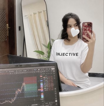 A woman with long brown hair wearing a white t-shirt is standing in front of a mirror holding her phone, taking a selfie. She appears happy as she poses for the photo. The mirror reflects the image of the bathroom sink that contains a vase filled with pretty flowers. A computer monitor is visible on a table behind the woman. On the monitor screen, there are stock market graphs showing fluctuations in different sectors.