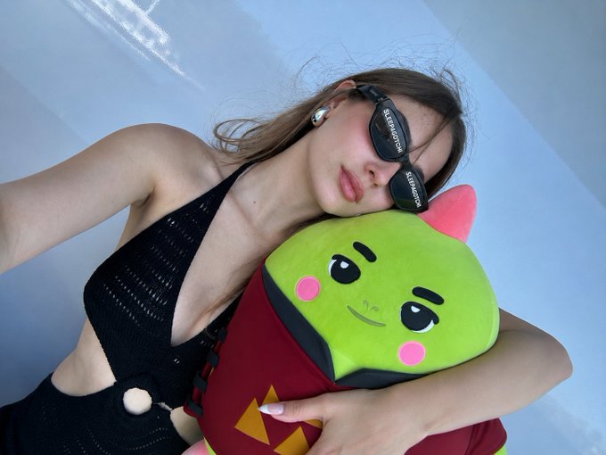 A woman wearing black Balenciaga sunglasses and a black cutout swimsuit rests her head on a green plush character toy.
