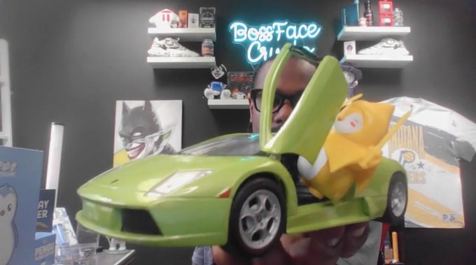 A collector's room displays various pop culture items including a Batman print and a 'Boss Face' neon sign, with someone holding a green toy Lamborghini with scissor doors and a yellow plush character inside.