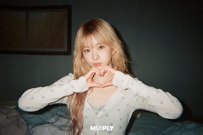 A blonde woman in a white top with purple hearts makes a heart shape with her hands, with the watermark 'MU:PLY' in the corner.
