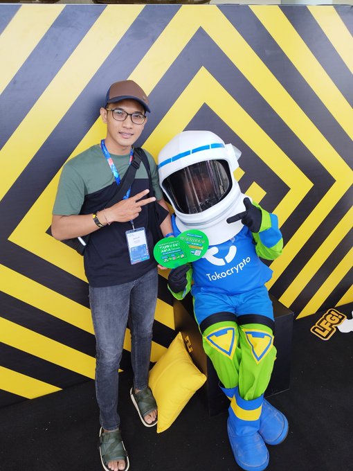A man in a green shirt and brown cap poses with a peace sign next to a person in a blue and green Tokocrypto mascot astronaut costume. They stand in front of a yellow and black geometric patterned wall, with the mascot holding a 'Tokocrypto' promotional sign.