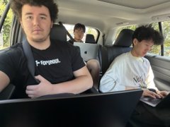 A candid in-car photo showing a bearded man in a black 'Torqeedo' t-shirt sitting in the front passenger seat with a laptop, while another young man works on a laptop in the back seat of a vehicle.