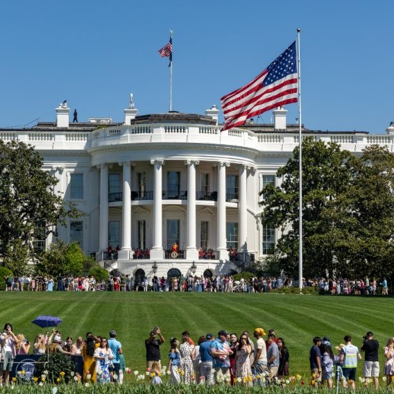 The White House stands prominently in the background as a large crowd gathers on the South Lawn under a clear blue sky, with the American flag waving from its iconic portico. The image complements the post by visually representing the center of federal power where the reported agreement on new crypto ethics rules—part of a revised 635-page bill aimed at saving the CLARITY Act—would be enforced on government officials.