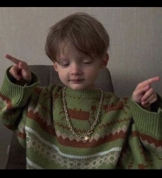 A young child with reddish-brown hair wearing a green patterned sweater and gold chain necklace, eyes closed with arms raised and hands crossed in a gesture.